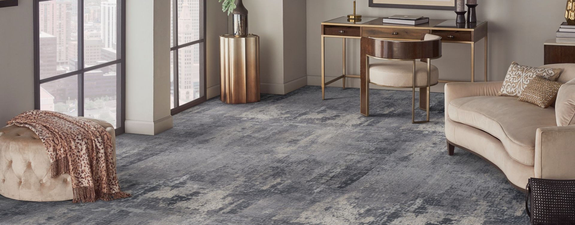 Contemporary living room with a grey and beige abstract carpet, gold-accented furniture, and a large window offering a view of tall buildings.